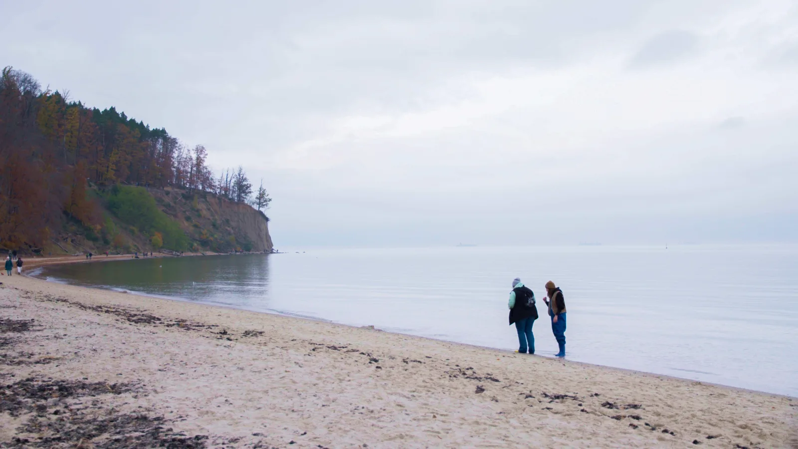 Dos personas en una playa de arena bajo el acantilado de Orłowo, superficie tranquila del Báltico en un día nublado