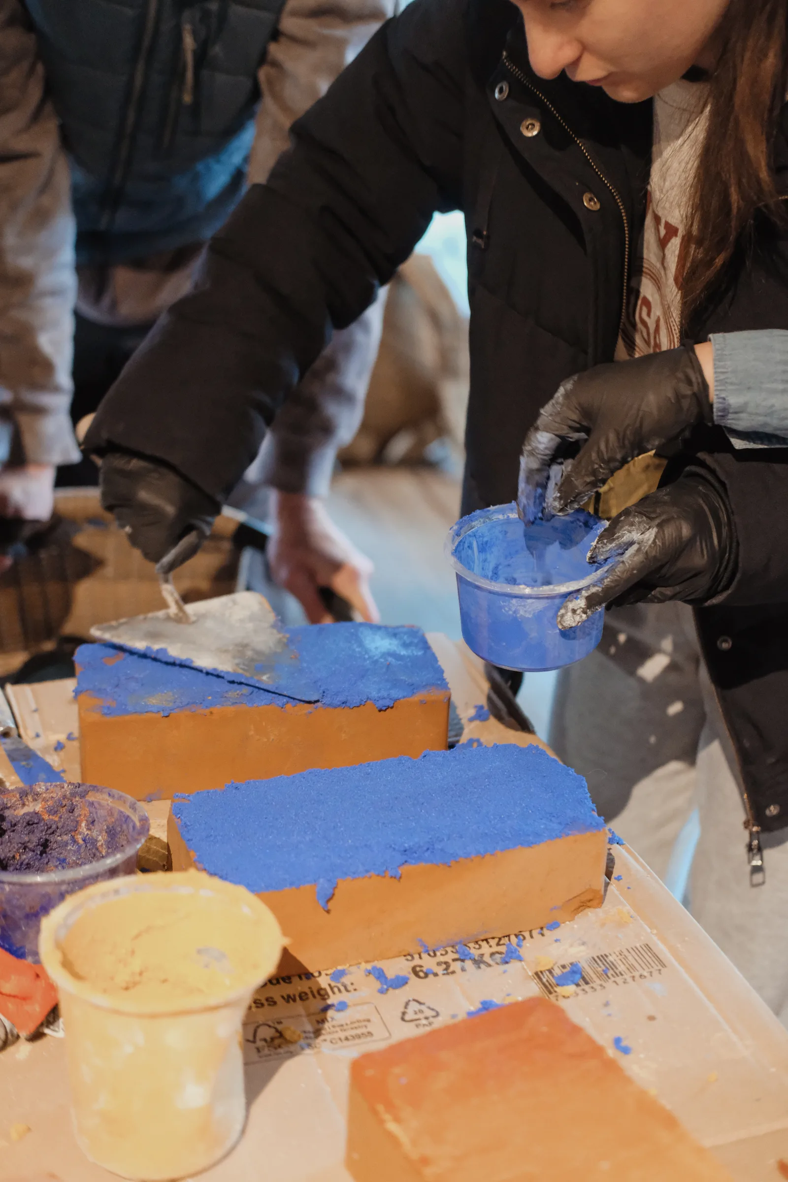 Manos con guantes aplican un revoque azul Shell White sobre bloques de ladrillo durante un taller